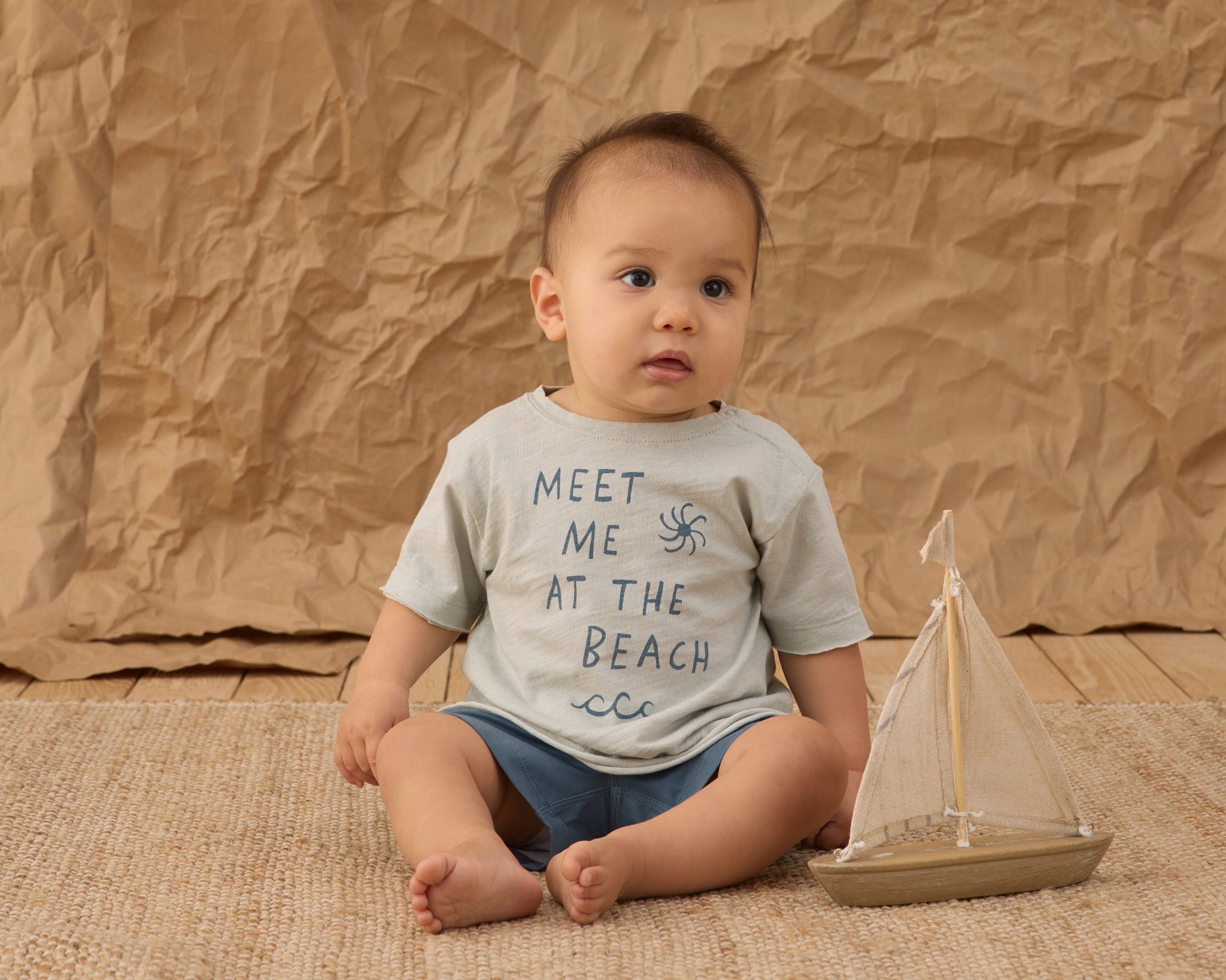 Baby sitting on a jute rug wearing the Rylee & Cru Paneled Boardshort in Marine with a beach tee