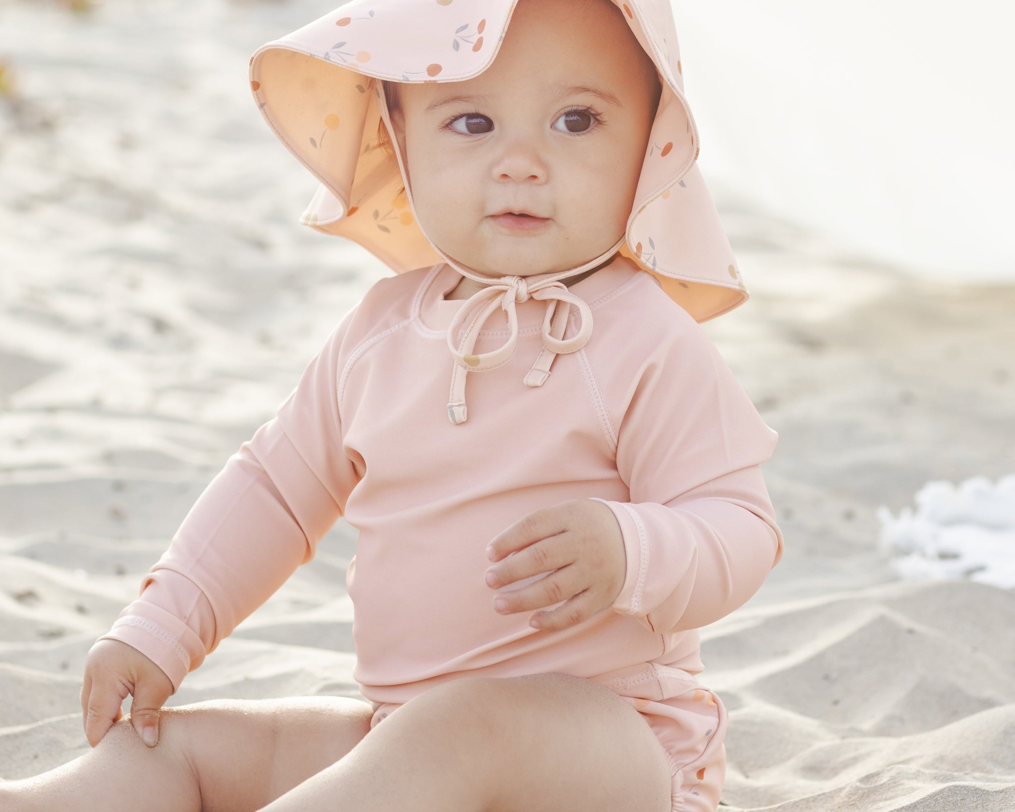 Baby in a floral sun bonnet sitting on the sand wearing the Rylee & Cru Rashguard Long Sleeve in Blush