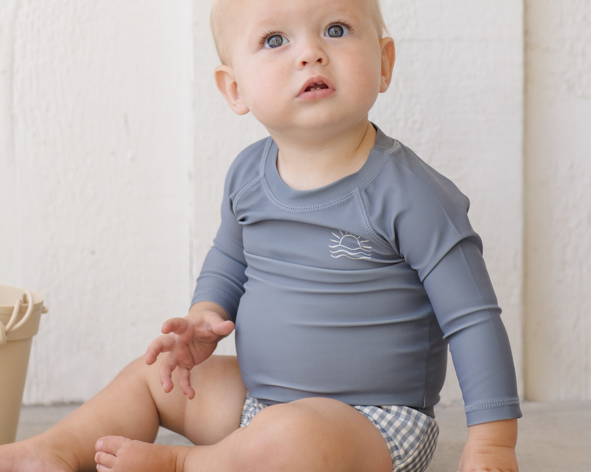 Baby sitting in the Rylee & Cru Rashguard Long Sleeve in Ocean with gingham swim trunks
