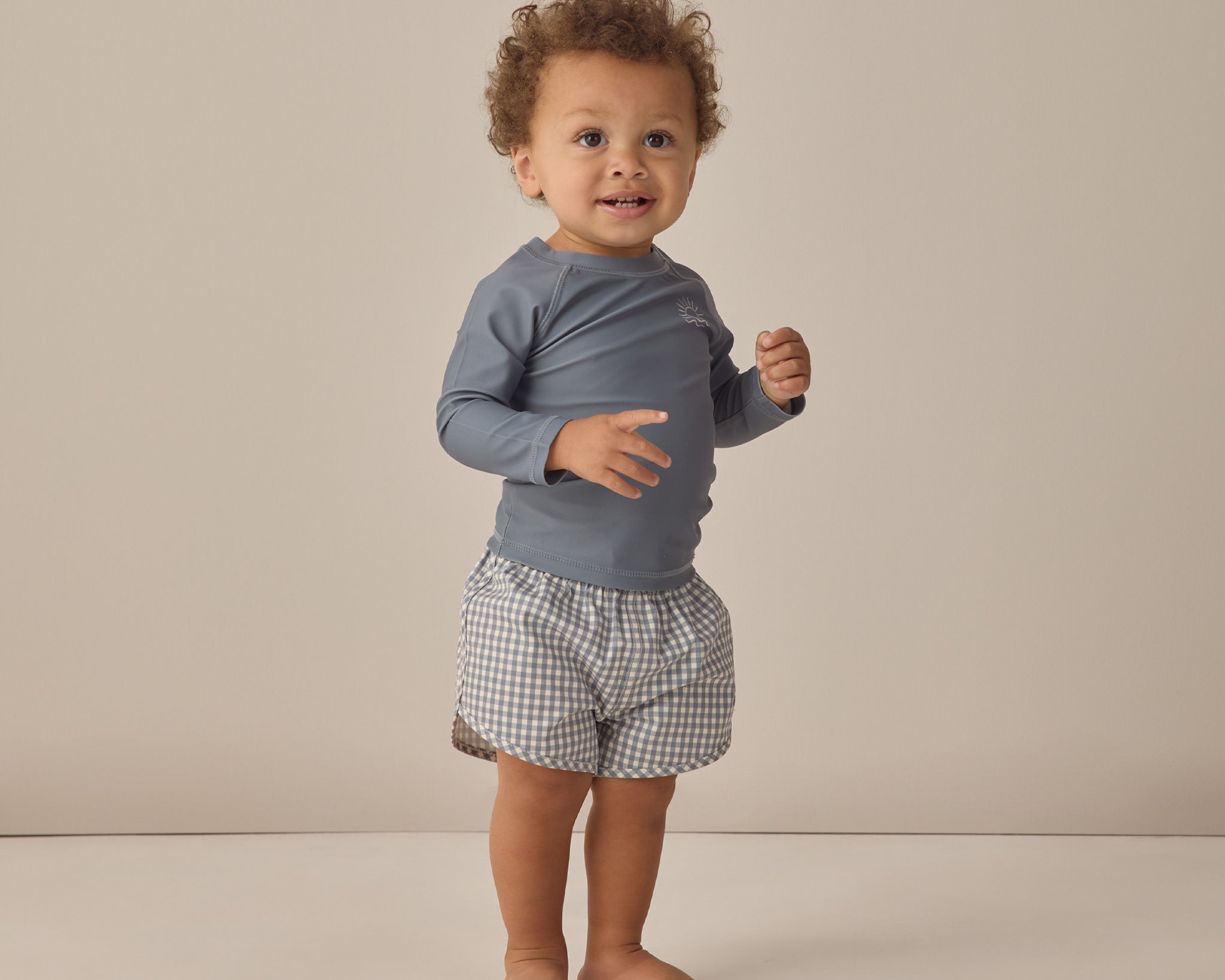 Smiling toddler standing in the Rylee & Cru Rashguard Long Sleeve in Ocean with gingham trunks