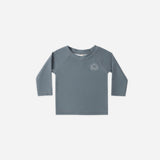Rylee & Cru Rashguard Long Sleeve in Ocean, a slate blue baby swim top with a small sun logo, laid flat
