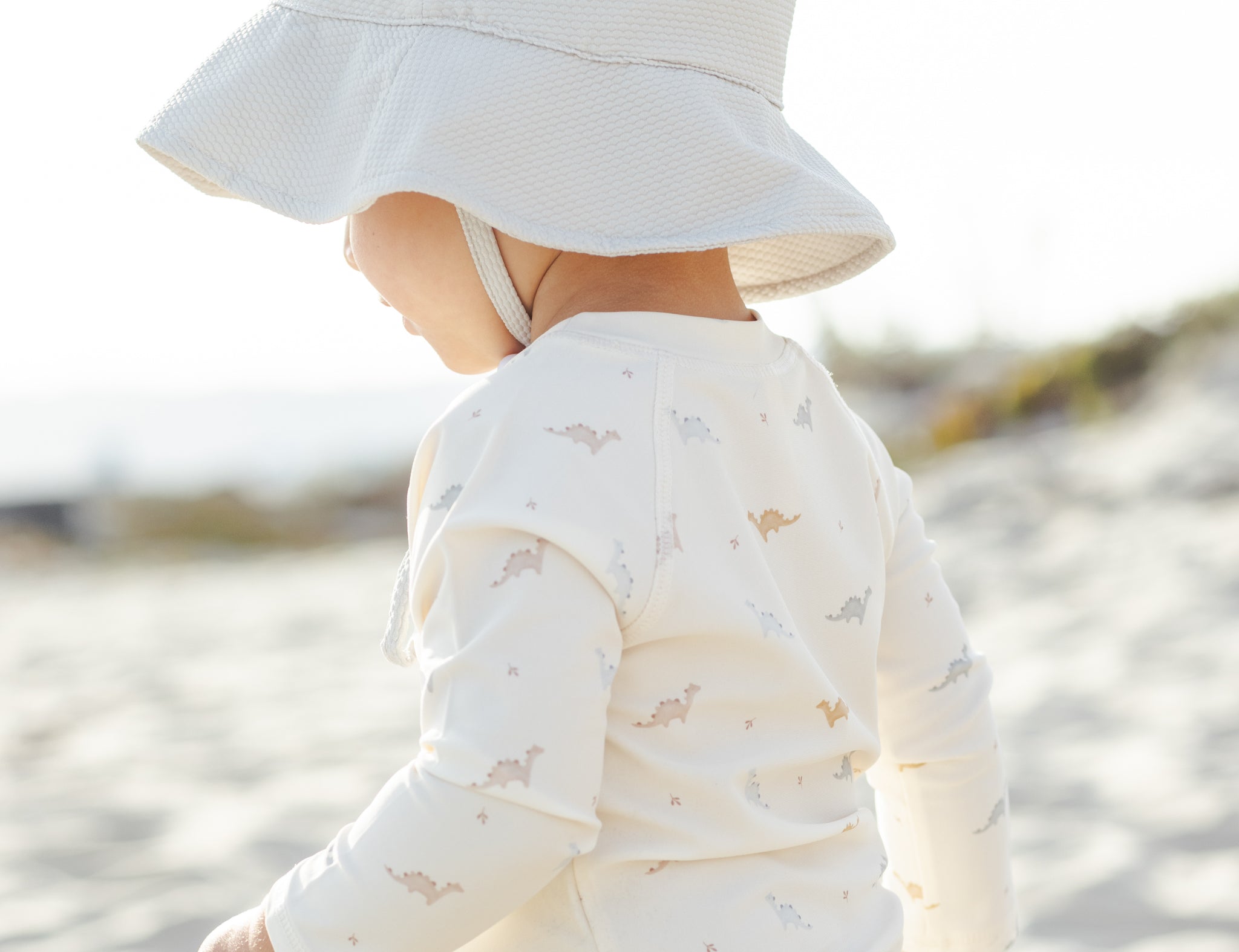 Toddler in a white sun hat wearing the Rylee & Cru Rashguard Top + Short Set in Dino at the beach, back view