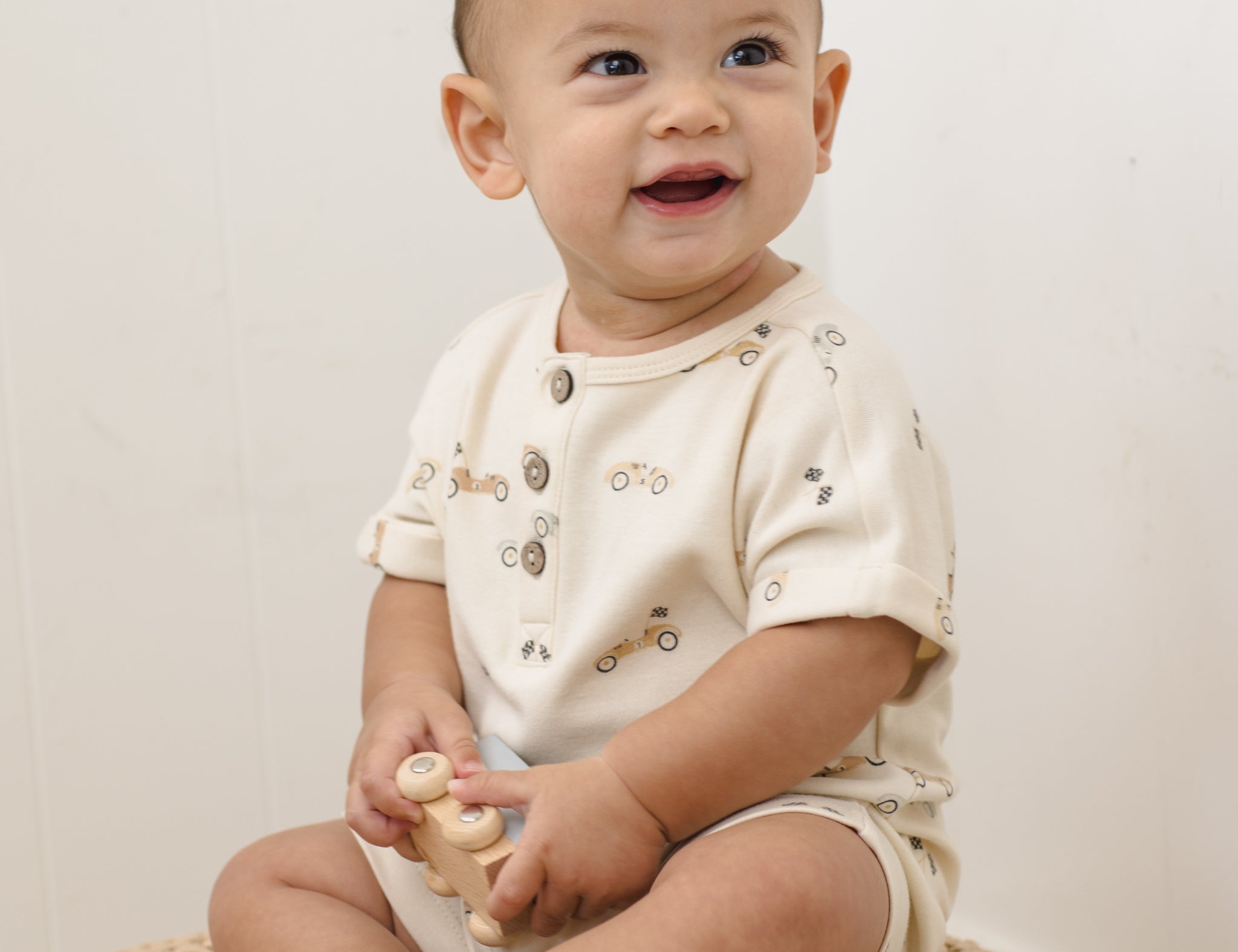 Smiling baby sitting on a jute rug wearing the Rylee & Cru Short Sleeve One-Piece in Race Car