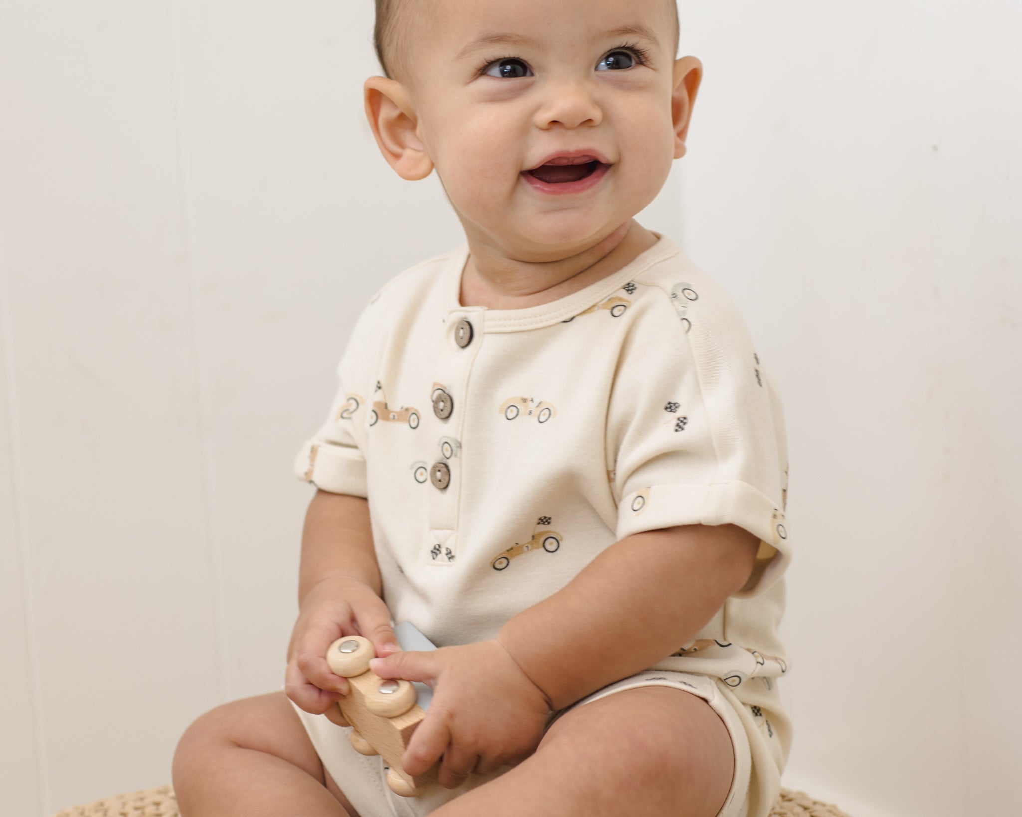 Smiling baby sitting on a jute rug wearing the Rylee & Cru Short Sleeve One-Piece in Race Car