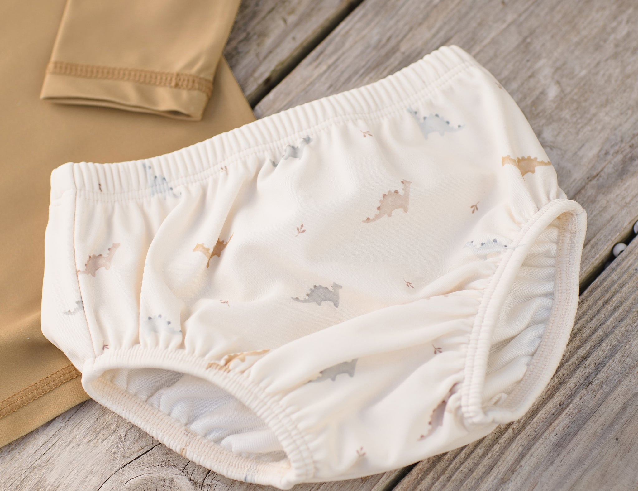 Rylee & Cru Swim Diaper in Dino laid on a wooden surface beside a tan rashguard