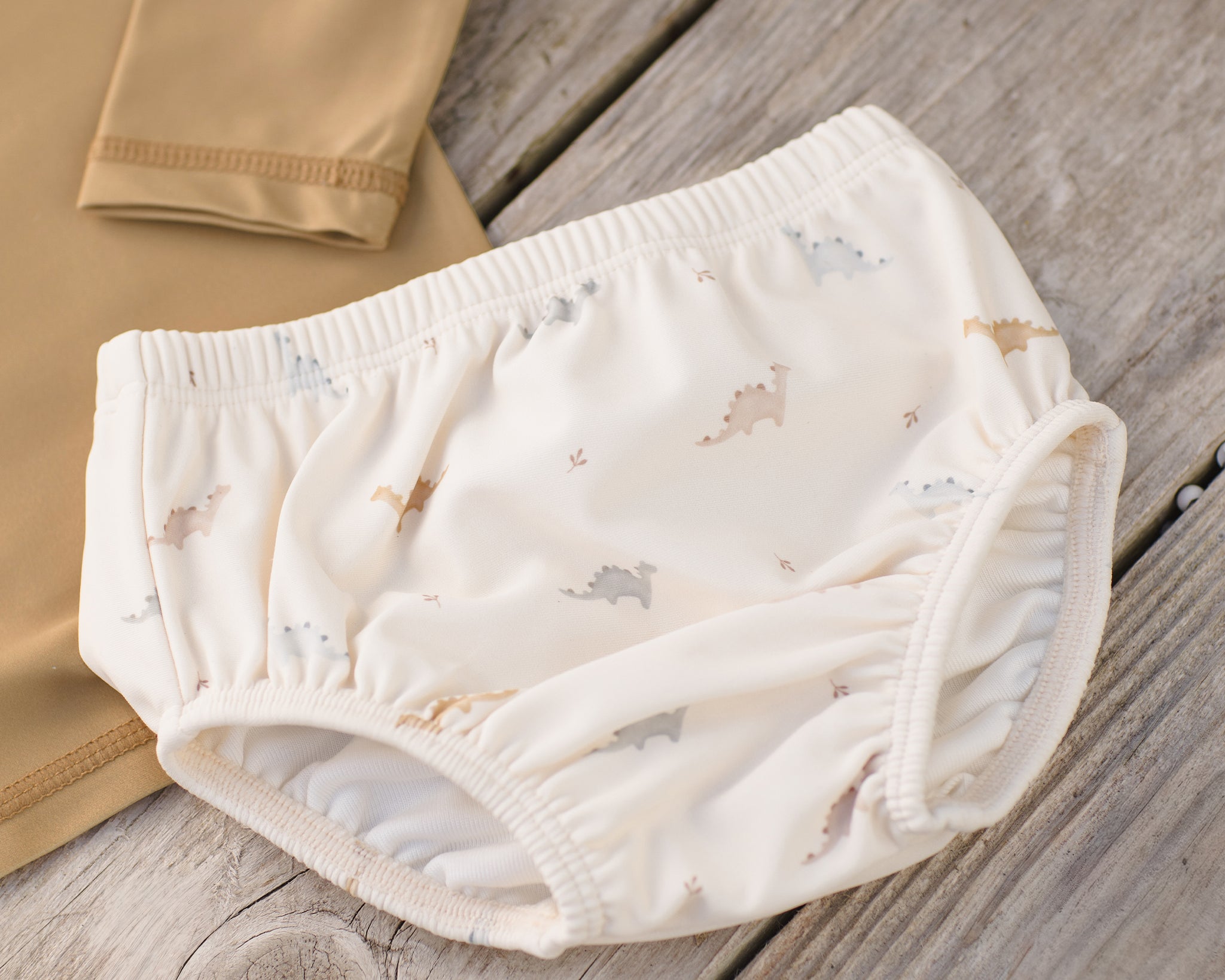 Rylee & Cru Swim Diaper in Dino laid on a wooden surface beside a tan rashguard