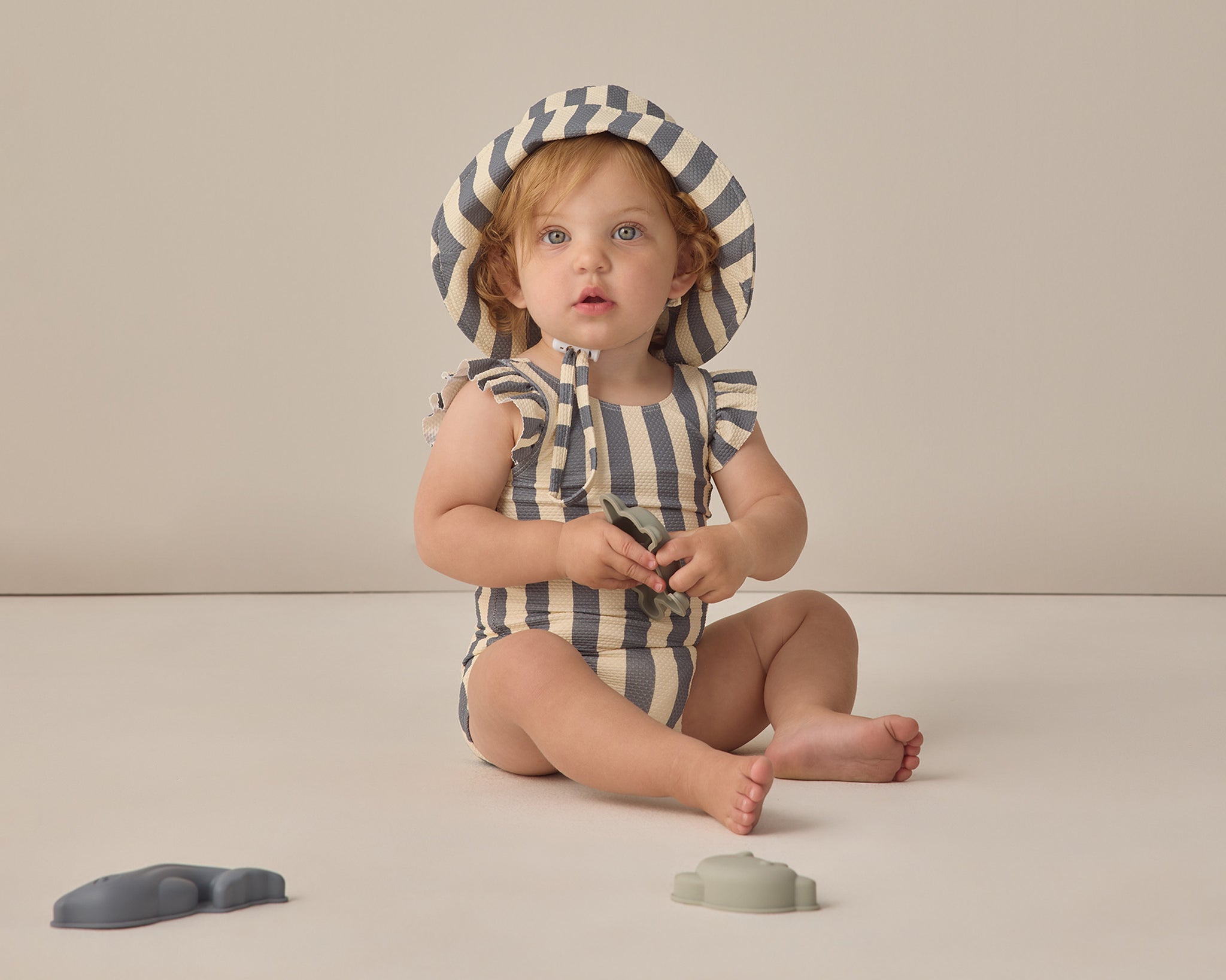 Baby sitting in a striped swimsuit wearing the Rylee & Cru Youth Bucket Hat in Retro Stripe Ocean