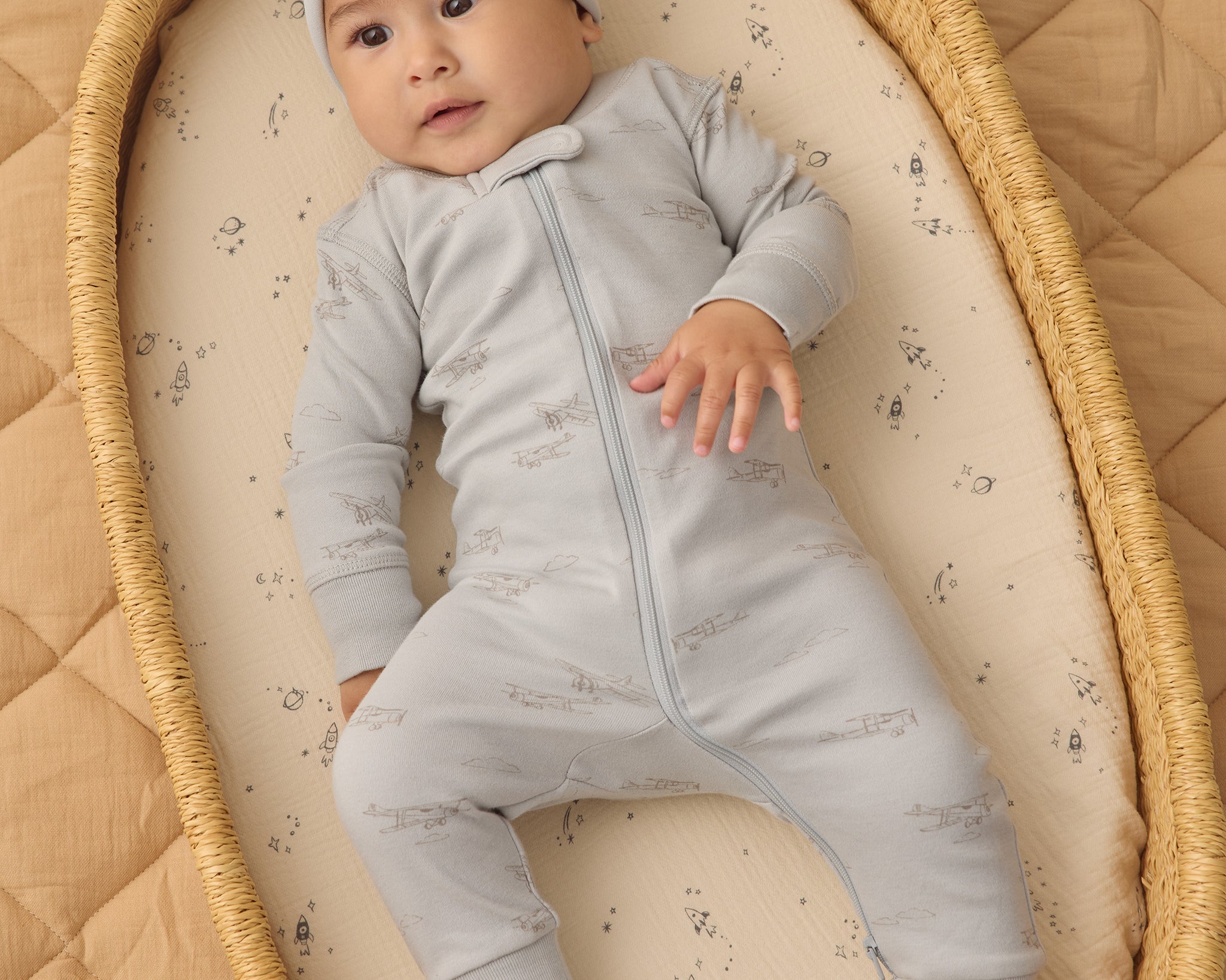 Baby in a bassinet wearing the Rylee & Cru Zip Longsleeve Sleeper in Airplane, a light blue sleeper with a matching hat