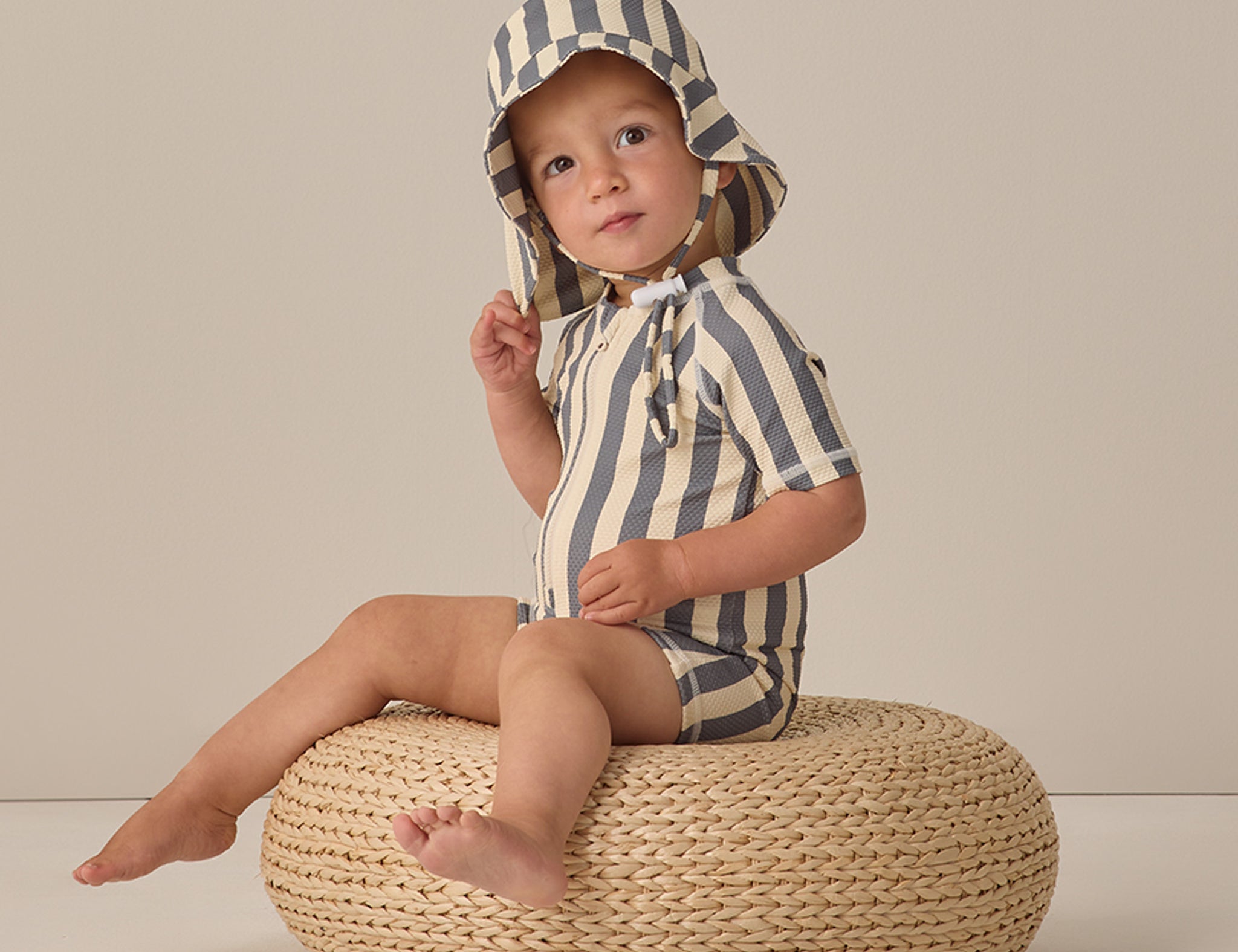 Toddler sitting on a wicker pouf wearing the Rylee & Cru Zip Rashguard One-Piece in Retro Ocean Stripe with a matching hat
