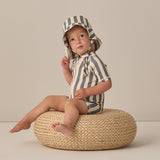 Toddler sitting on a wicker pouf wearing the Rylee & Cru Zip Rashguard One-Piece in Retro Ocean Stripe with a matching hat