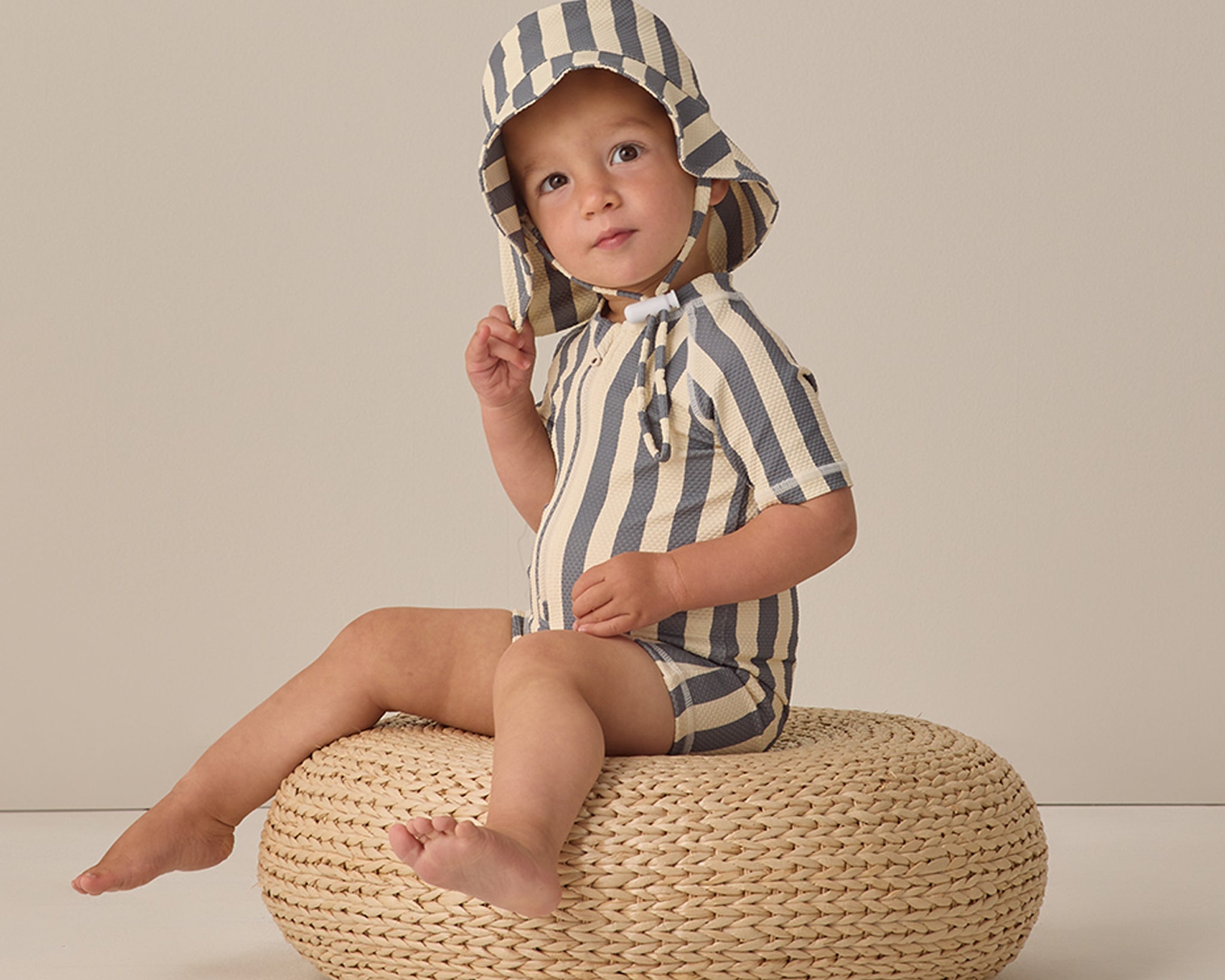 Toddler sitting on a wicker pouf wearing the Rylee & Cru Zip Rashguard One-Piece in Retro Ocean Stripe with a matching hat
