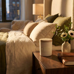 Skeem Black Currant Candle on a wooden nightstand beside a bed with white and olive bedding