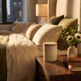 Skeem Black Currant Candle on a wooden nightstand beside a bed with white and olive bedding