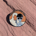 Round Take Me To Your Dirt enamel pin with a gray alien in sunglasses making a peace sign, on red sandstone in Moab, Utah.