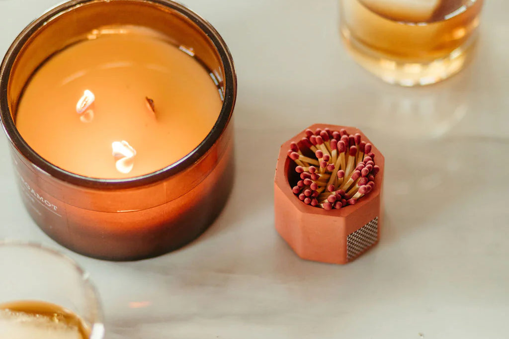 Tenn Prairie Concrete Match Holder in brick orange beside a lit amber candle and a glass of whiskey