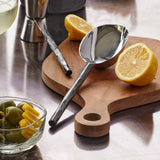 Texxture Chiku Teak Cutting Board on a counter with lemons, a spoon and a bowl of olives