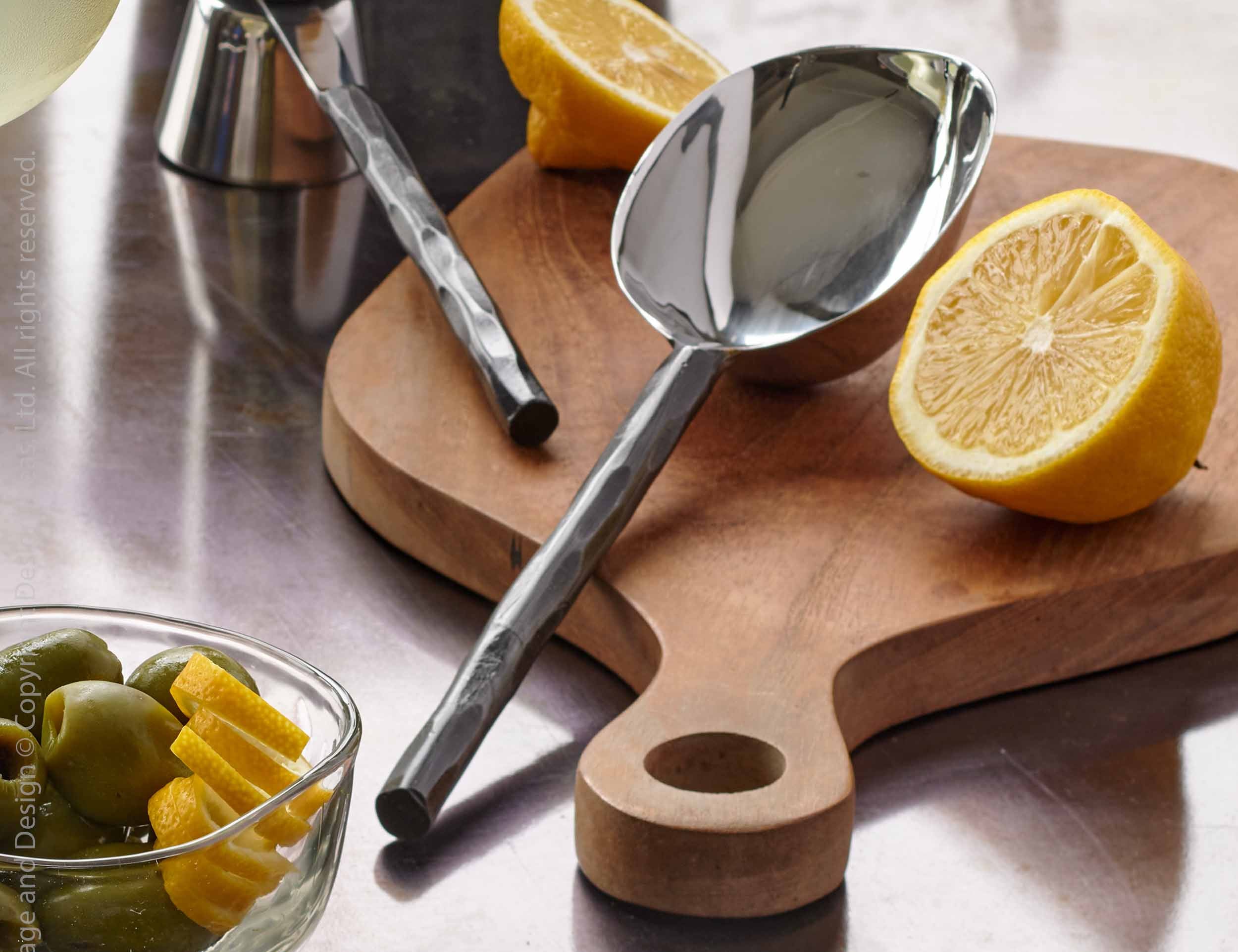 Texxture Chiku Teak Cutting Board on a counter with lemons, a spoon and a bowl of olives