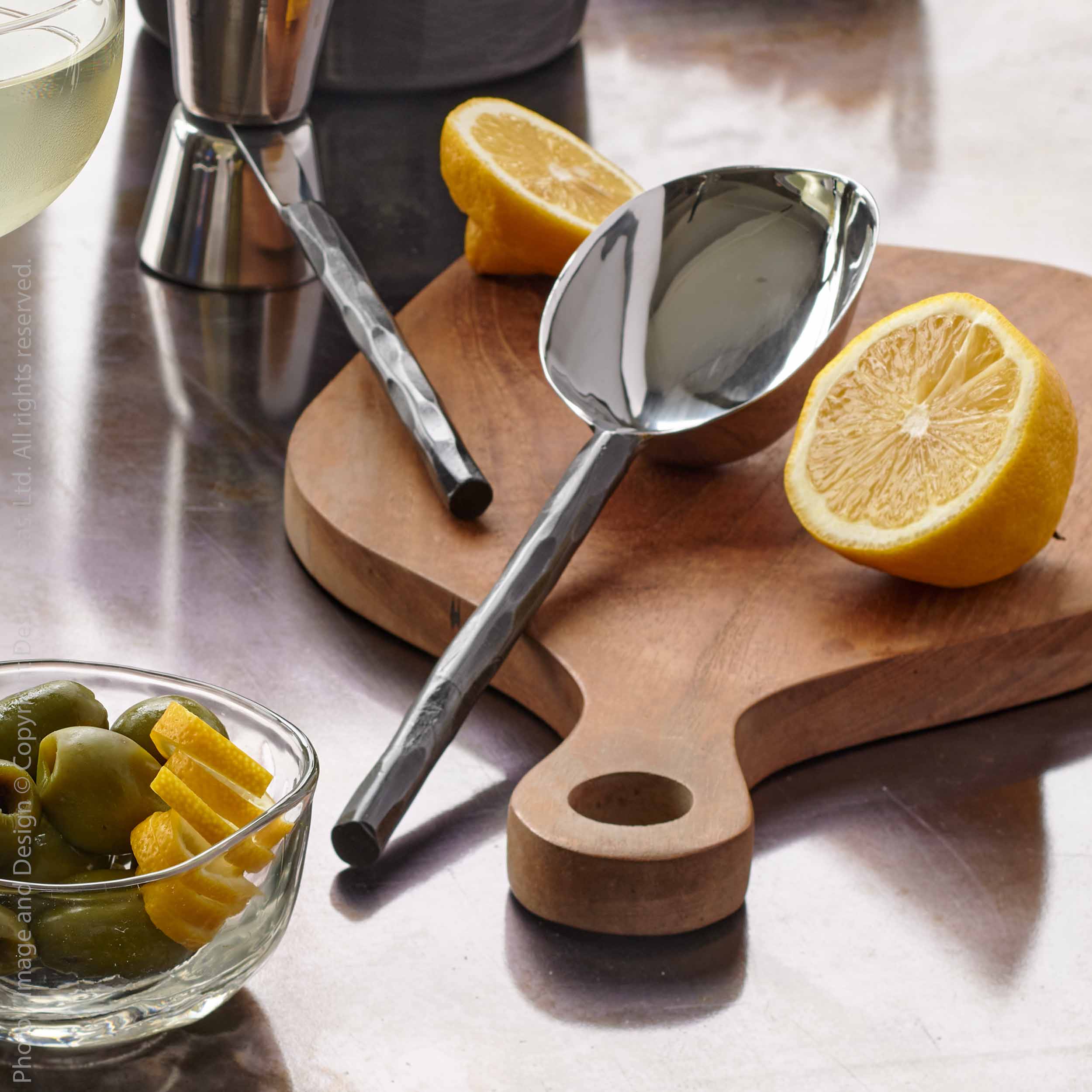 Texxture Chiku Teak Cutting Board on a counter with lemons, a spoon and a bowl of olives