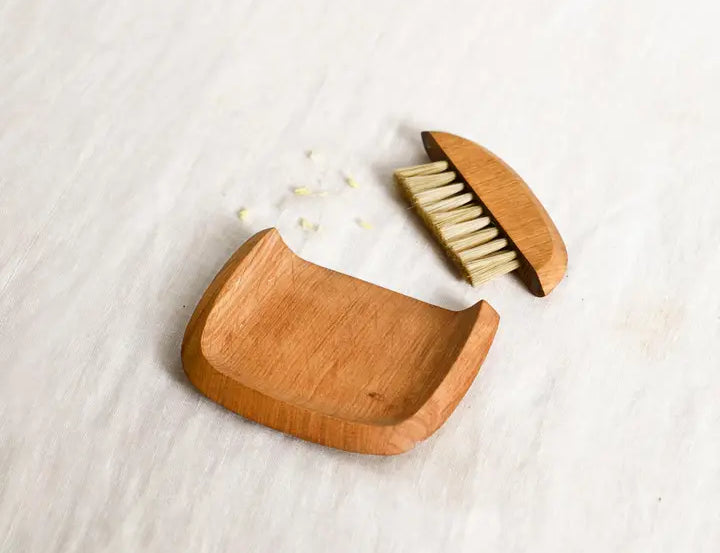 The Collective Belfort Crumb Brush Set, a natural wood dustpan and bristle brush on white linen