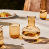 The Collective Ember Carafe + Tumbler Set in amber glass side by side on an outdoor dining table