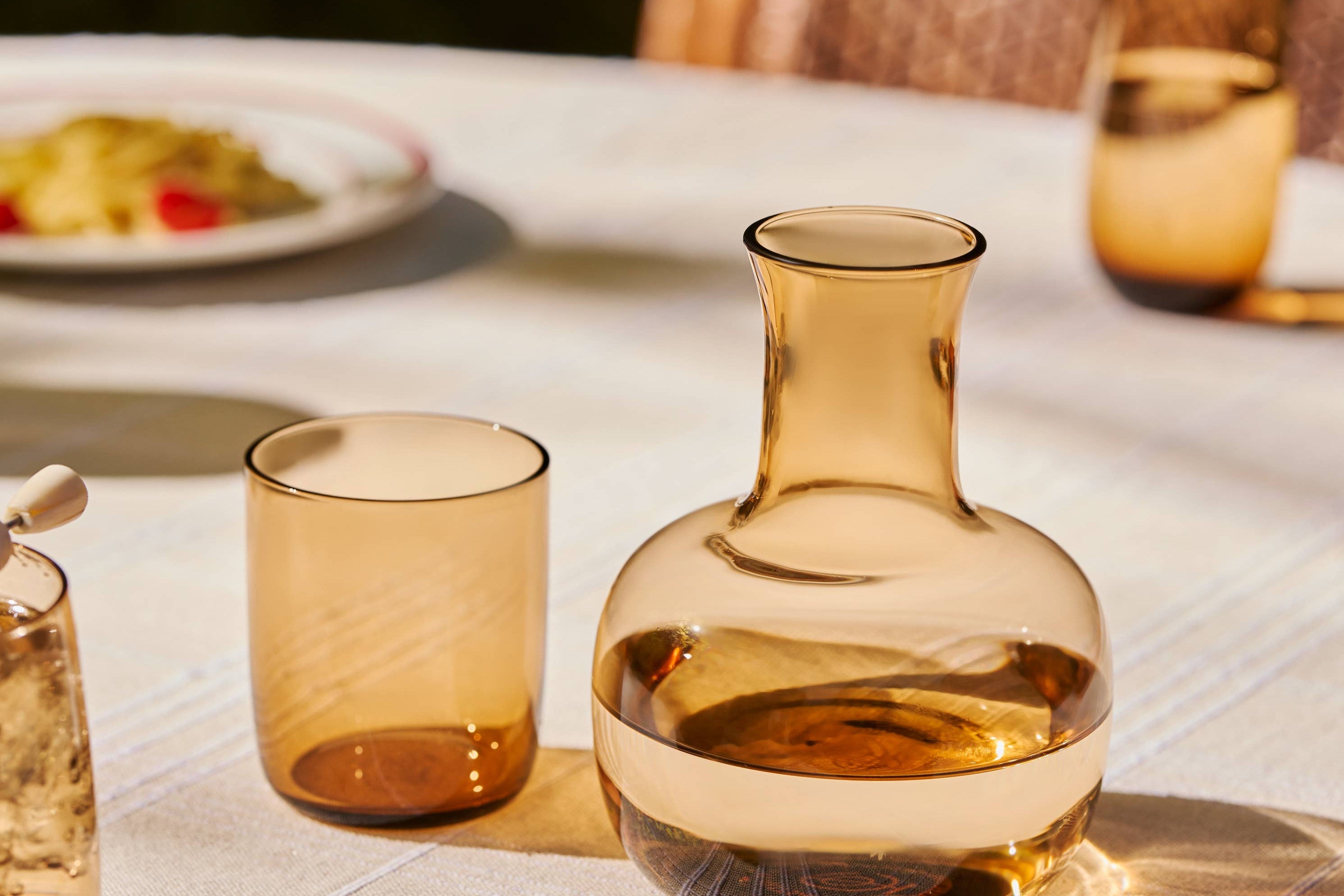 The Collective Ember Carafe + Tumbler Set in amber glass side by side on an outdoor dining table