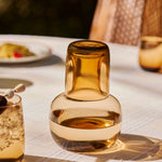 The Collective Ember Carafe + Tumbler Set, amber glass carafe with the tumbler stacked on top, on an outdoor table