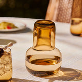 The Collective Ember Carafe + Tumbler Set, amber glass carafe with the tumbler stacked on top, on an outdoor table