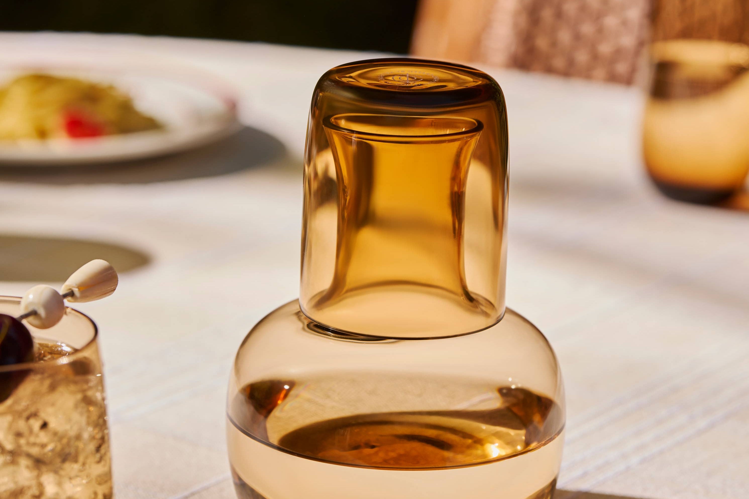 The Collective Ember Carafe + Tumbler Set, amber glass carafe with the tumbler stacked on top, on an outdoor table