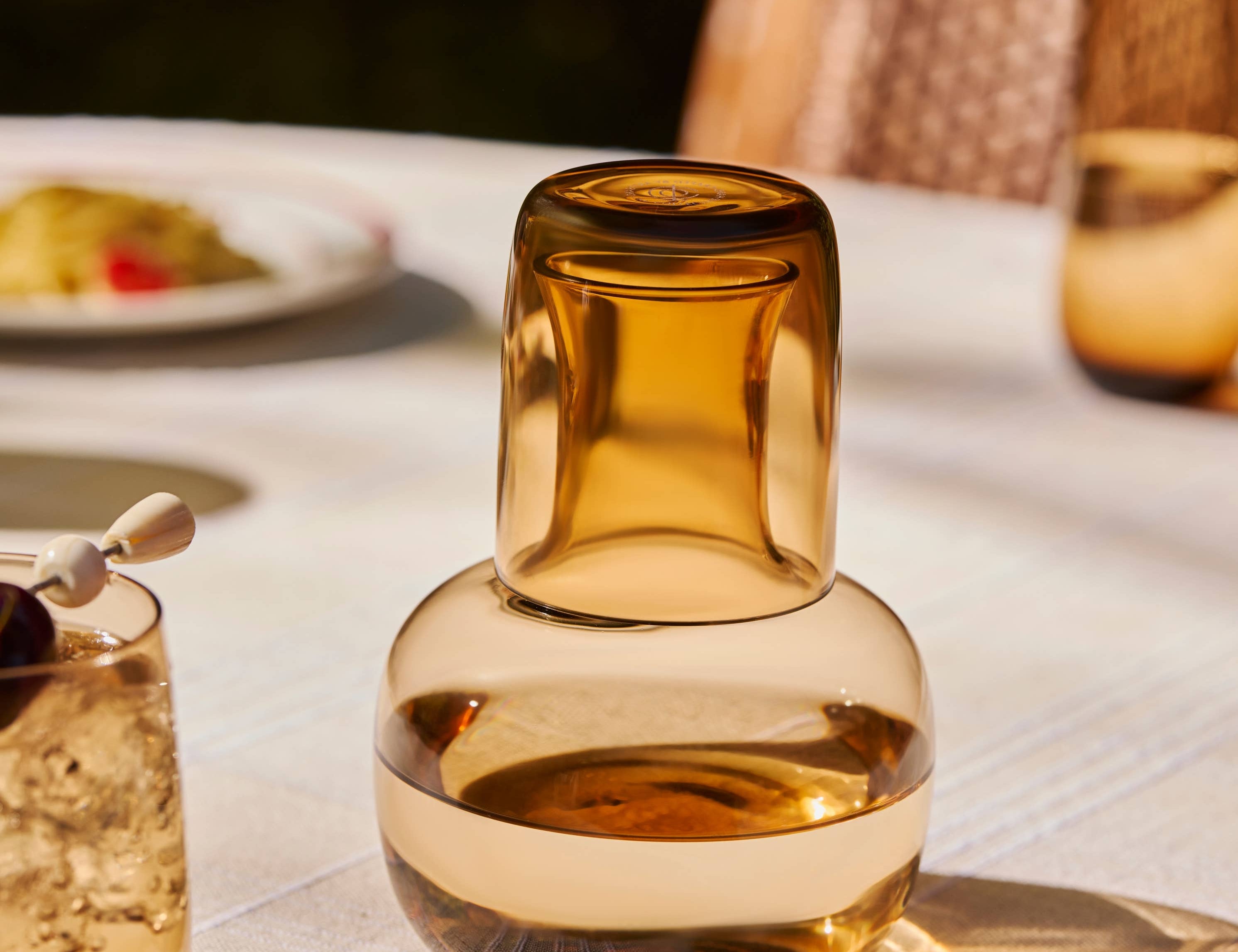 The Collective Ember Carafe + Tumbler Set, amber glass carafe with the tumbler stacked on top, on an outdoor table