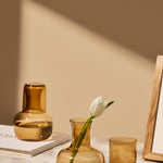 The Collective Ember Carafe + Tumbler Set in amber glass on a table with a tulip in a vase