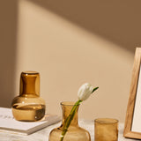 The Collective Ember Carafe + Tumbler Set in amber glass on a table with a tulip in a vase