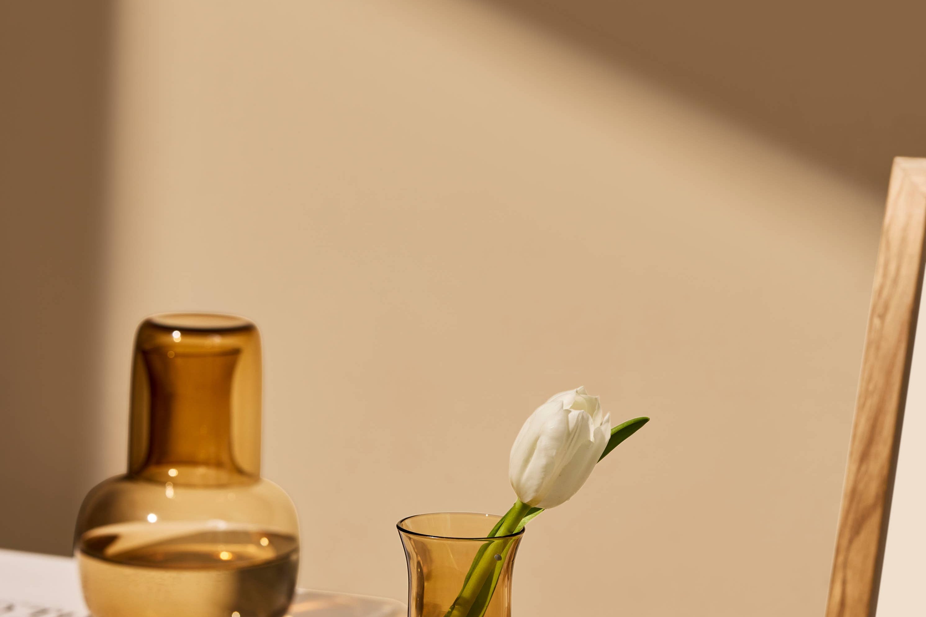 The Collective Ember Carafe + Tumbler Set in amber glass on a table with a tulip in a vase