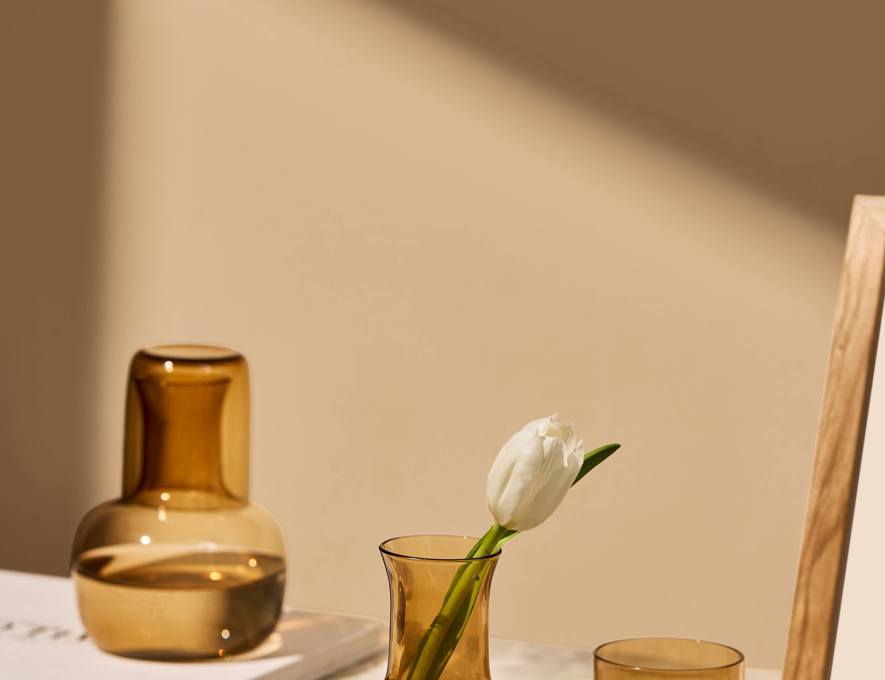 The Collective Ember Carafe + Tumbler Set in amber glass on a table with a tulip in a vase