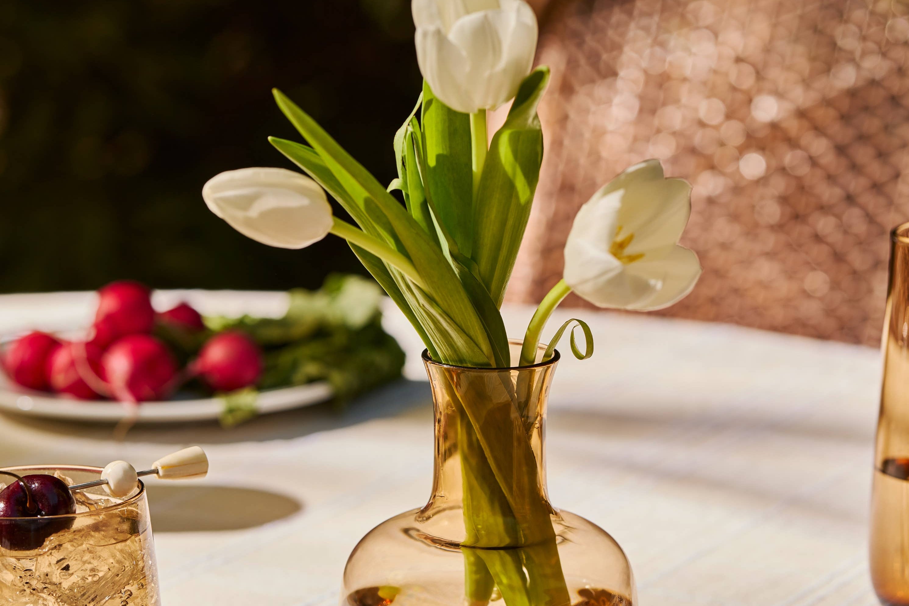 The Collective Ember amber glass carafe holding white tulips on a table set with radishes
