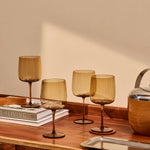 The Collective Ember Wine Glasses Set of 4 in amber glass on a wooden table beside books