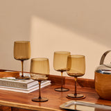 The Collective Ember Wine Glasses Set of 4 in amber glass on a wooden table beside books