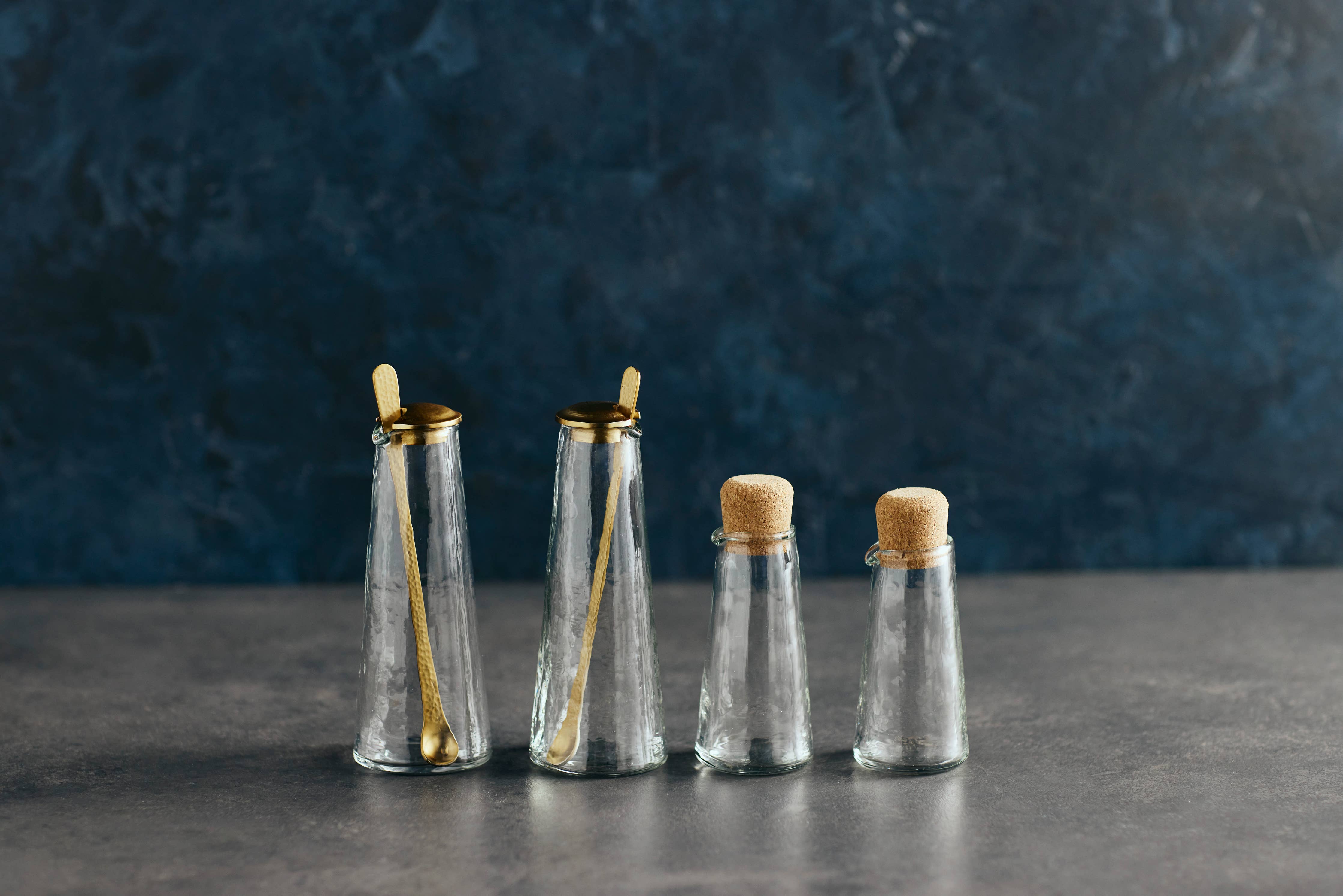 The Collective Pebbled Small Carafes and shakers with cork stoppers and gold spoons on a dark surface