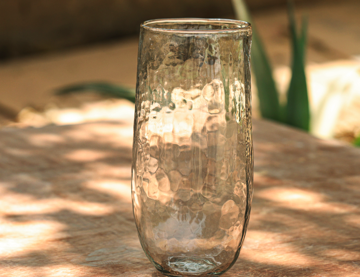The Collective Pebbled Tall Vase, a slim textured glass vase on a wooden table in dappled light