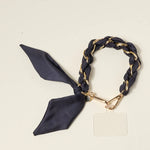 The Darling Effect Phone Scarf in Black, a satin scarf braided through a gold chain wristlet with a phone tether tab