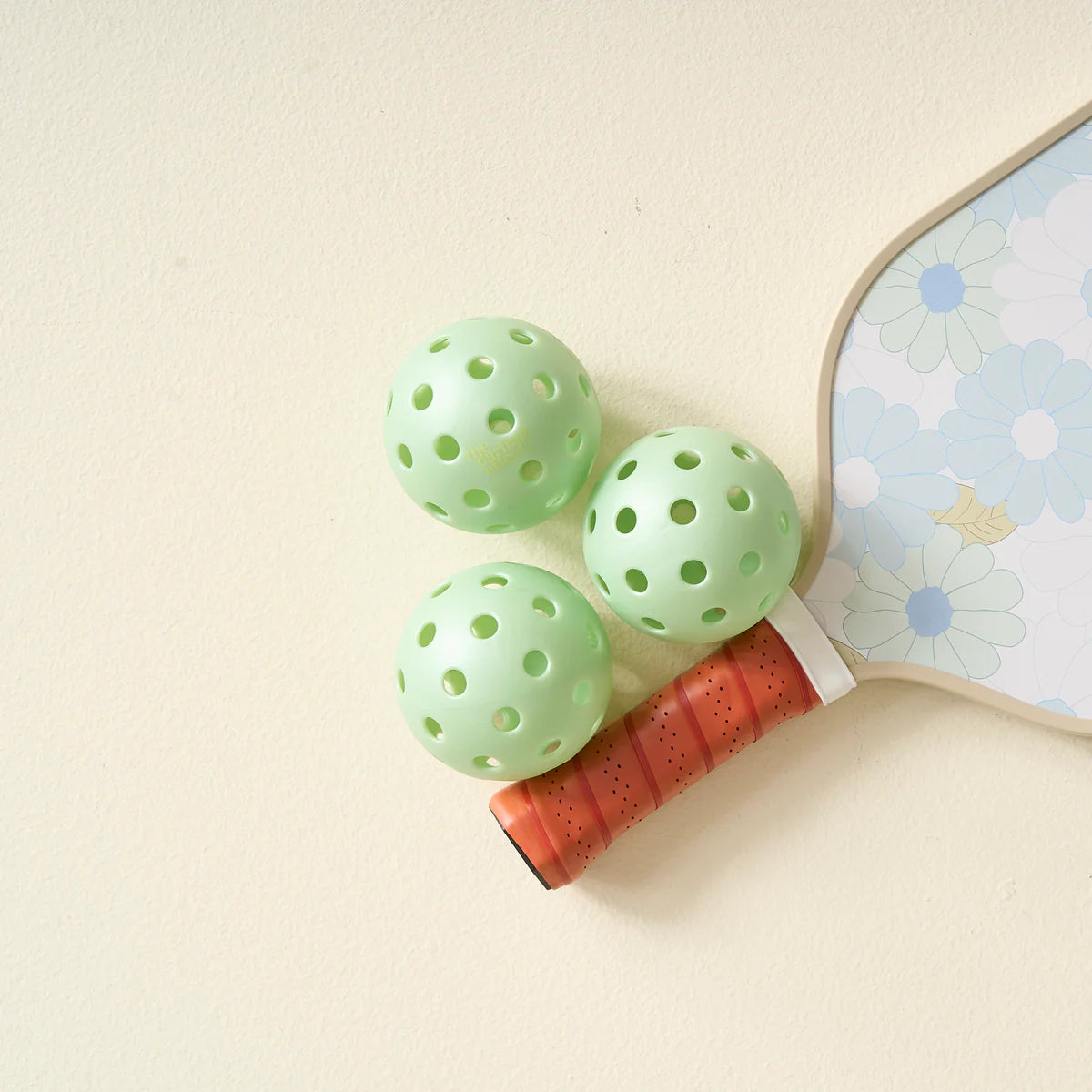 Three aqua pickleballs from The Darling Effect Pickleball Ball Set beside a daisy paddle