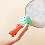 Hand holding a daisy paddle and an aqua pickleball from The Darling Effect Pickleball Ball Set