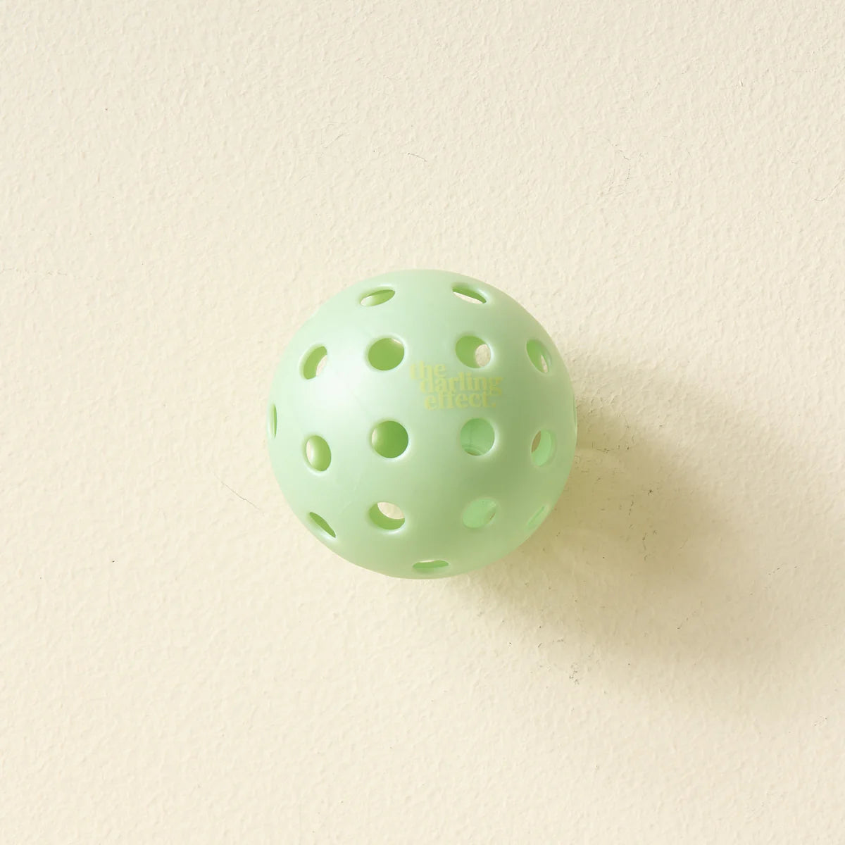 Single mint green pickleball from The Darling Effect Pickleball Ball Set on a cream background