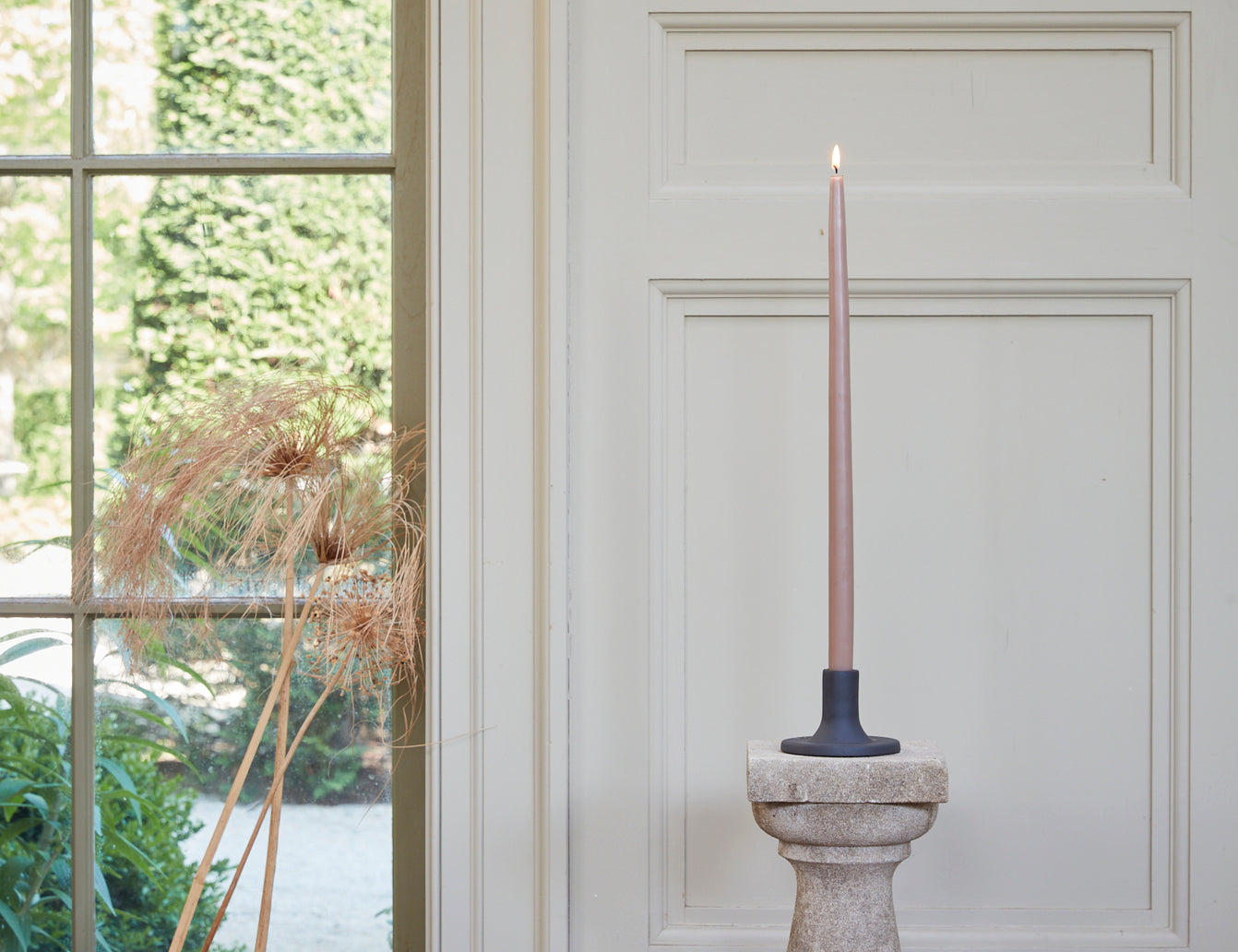 The Floral Society 18-inch Dipped Taper Candle in a muted rose shade, lit in a stone candlestick in front of a bright window