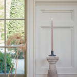 The Floral Society 18-inch Dipped Taper Candle in a muted rose shade, lit in a stone candlestick in front of a bright window