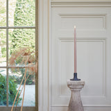 The Floral Society 18-inch Dipped Taper Candle in a muted rose shade, lit in a stone candlestick in front of a bright window