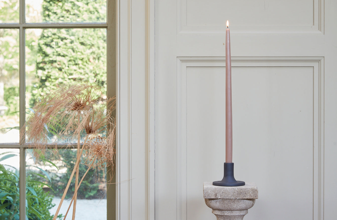 The Floral Society 18-inch Dipped Taper Candle in a muted rose shade, lit in a stone candlestick in front of a bright window