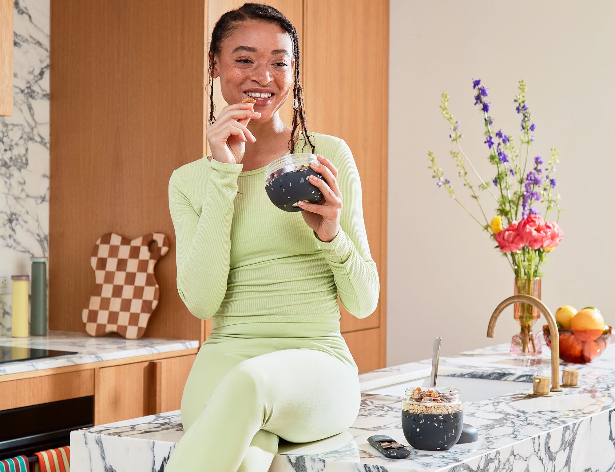 Smiling woman in green eating from a W&P Seal Tight Bowl in Terrazzo Charcoal at a marble counter